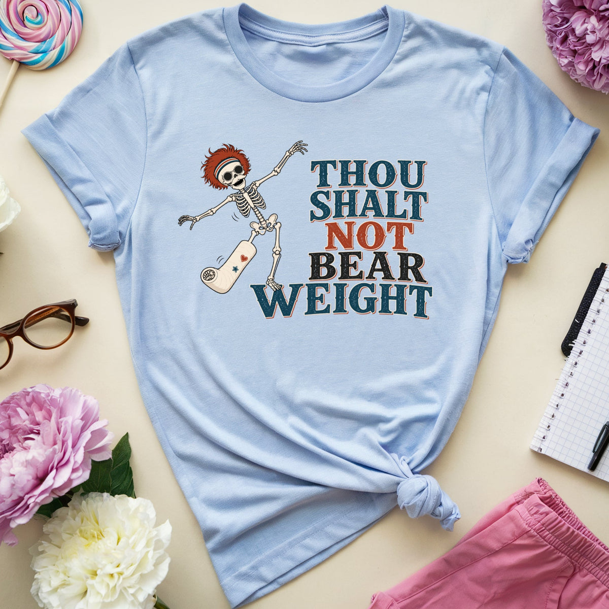A light blue graphic t-shirt with a distressed print of a skeleton and the text 'THOU SHALT NOT BEAR WEIGHT' in dark teal, orange, and brown. The shirt is styled in a flat-lay arrangement on a beige background with pink and white peonies, glasses, a lollipop, a notebook, and pink shorts.