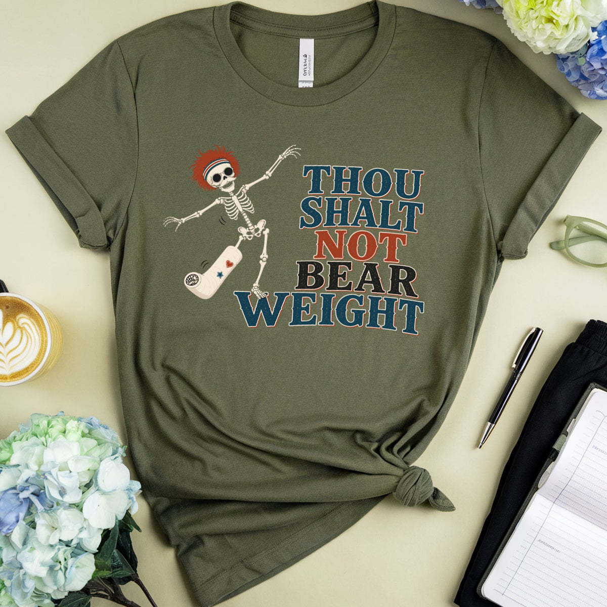 An olive green graphic t-shirt featuring a skeleton dancing on a skateboard with the text 'THOU SHALT NOT BEAR WEIGHT' in blue, red, and black. The shirt is styled in a flat-lay arrangement with a coffee cup, flowers, eyeglasses, a pen, and an open notebook on a light beige background.