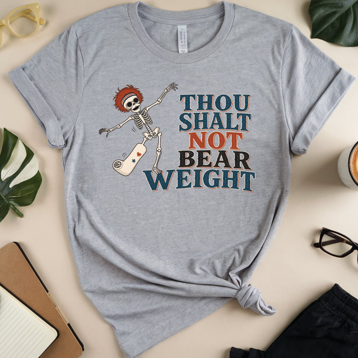 A heather gray graphic t-shirt laid flat on a beige surface, styled with a notebook, glasses, a coffee cup, and plant leaves. The t-shirt features a cartoon skeleton with red hair and the text 'THOU SHALT NOT BEAR WEIGHT' in blue, orange, and brown lettering.