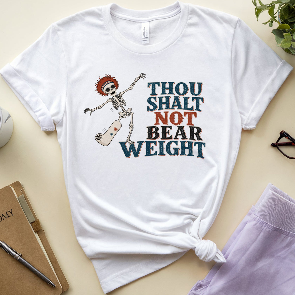 A white graphic t-shirt laid flat on a beige surface, styled with a coffee mug, notebook, glasses, and lavender pants. The t-shirt features a cartoon skeleton holding a skateboard next to the text 'THOU SHALT NOT BEAR WEIGHT' in dark blue and orange.