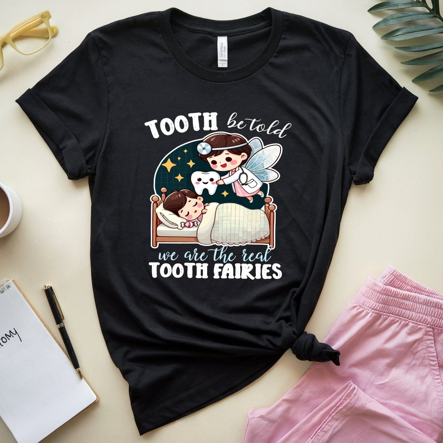 Black t-shirt with a graphic design of a tooth fairy and children, placed on a surface with a cup, notebook, and pink shorts.