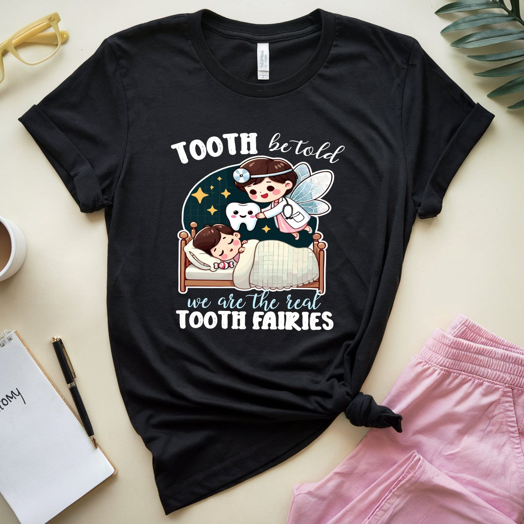 Black t-shirt with a graphic design of a tooth fairy and children, placed on a surface with a cup, notebook, and pink shorts.