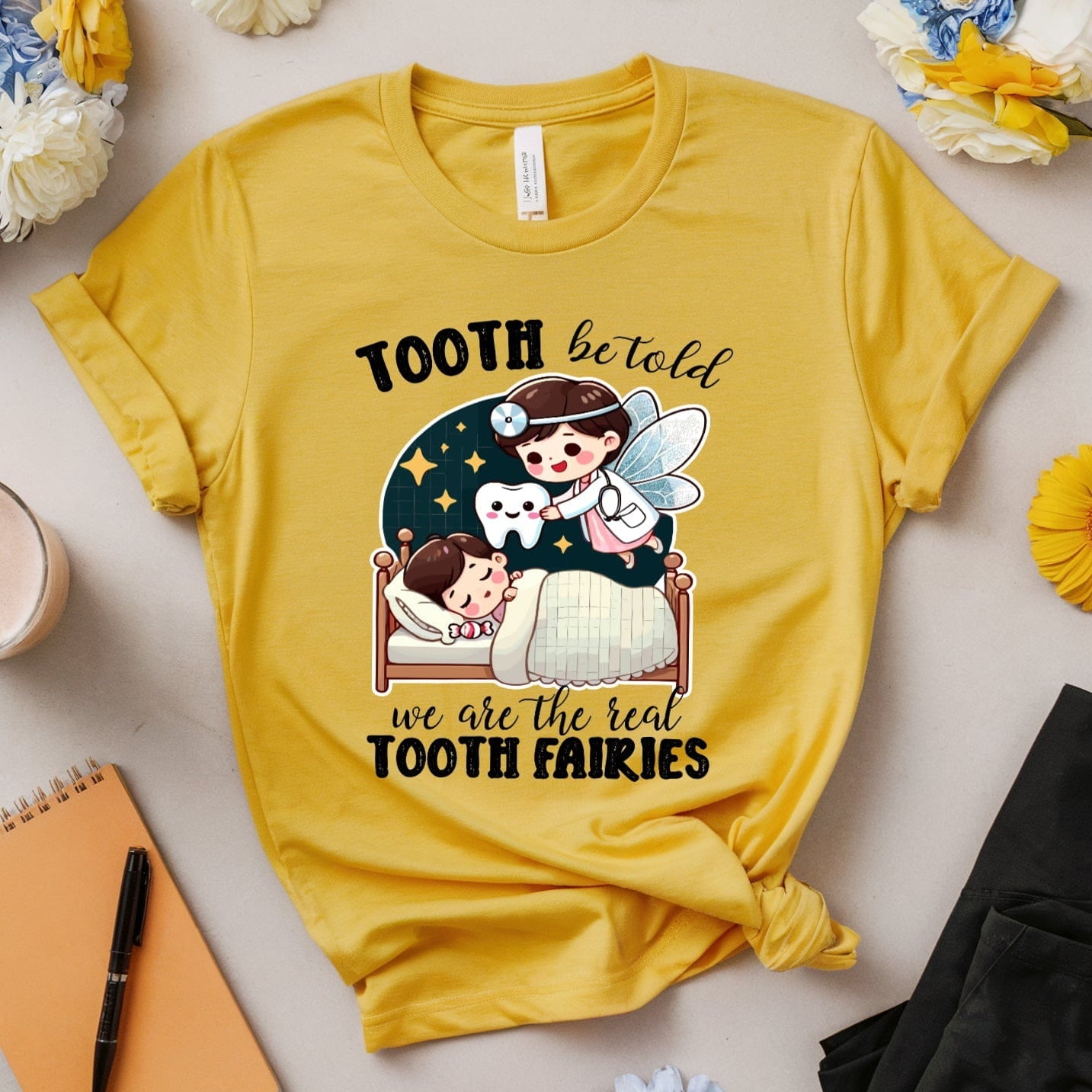 Yellow t-shirt with cartoon design and text, surrounded by flowers and accessories on a light surface.