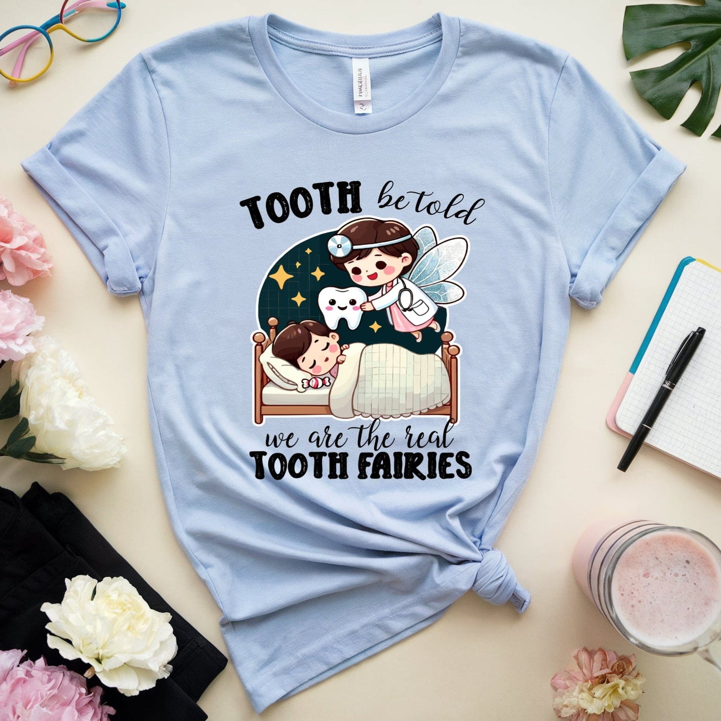 Light blue t-shirt with a graphic design of a tooth fairy and text, placed on a surface with flowers and a notebook.