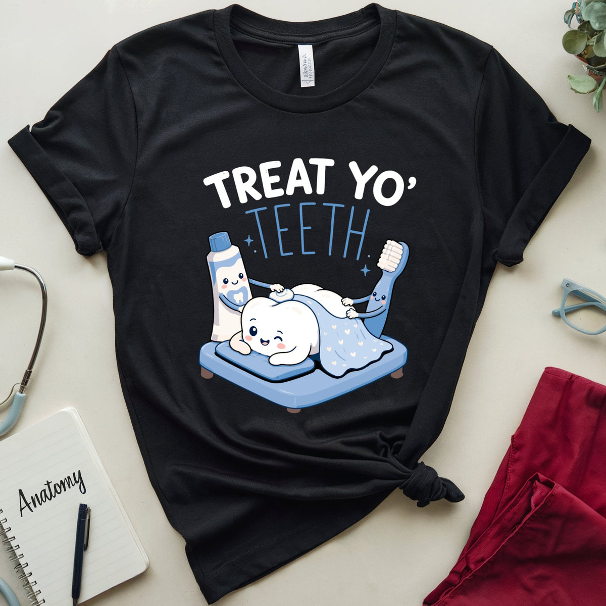 A black graphic T-shirt laid flat on a light beige surface, surrounded by dental-themed props including a stethoscope, notebook labeled 'Anatomy,' glasses, a plant, and red scrubs. The shirt has a cartoon of a white dog lying under a blue blanket with the text 'TREAT YO' TEETH.