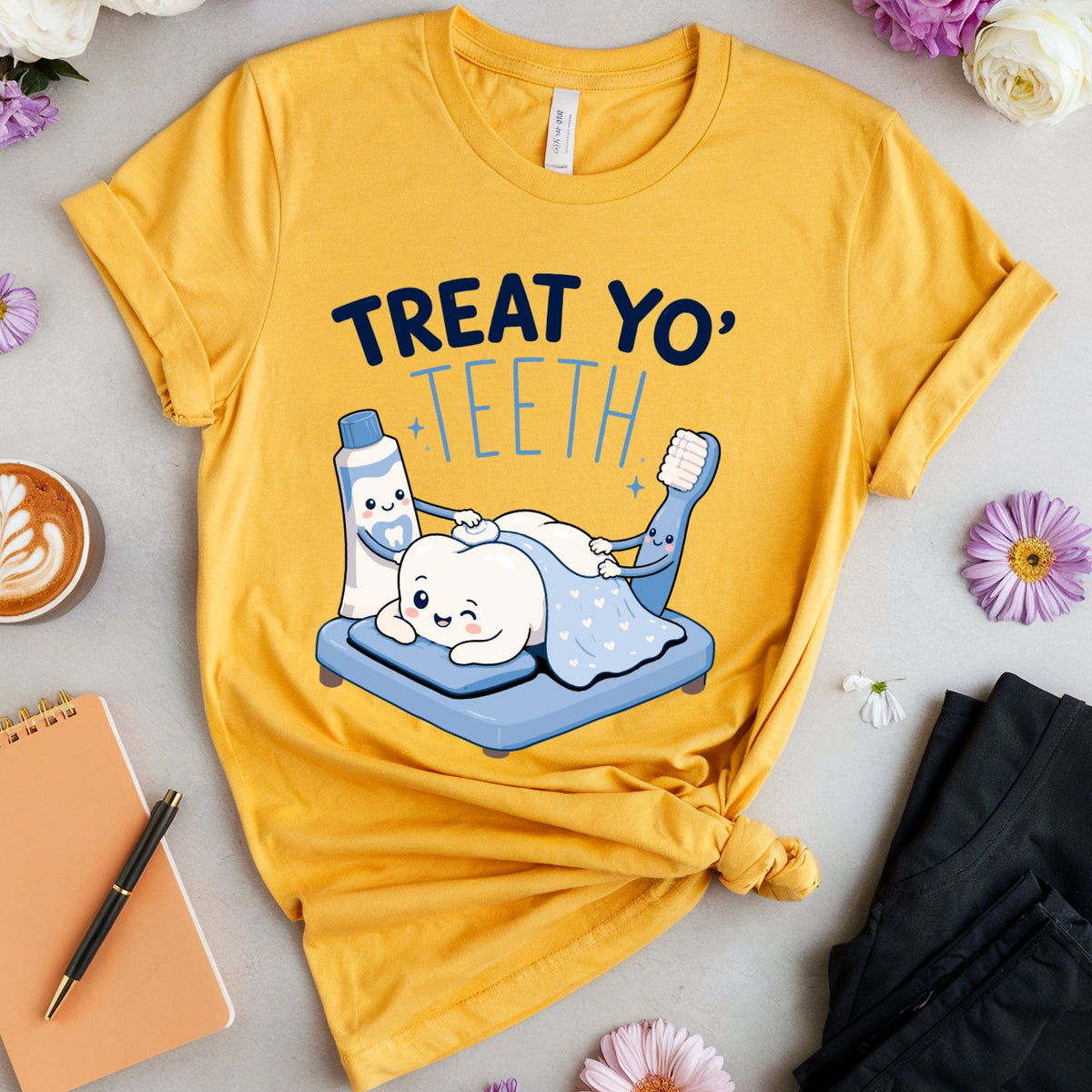 A yellow graphic T-shirt laid flat on a light gray surface, surrounded by flowers, a cup of latte, a notebook, a pen, and folded black pants. The shirt has a cartoon illustration of a tooth and the text 'TREAT YO' TEETH'.