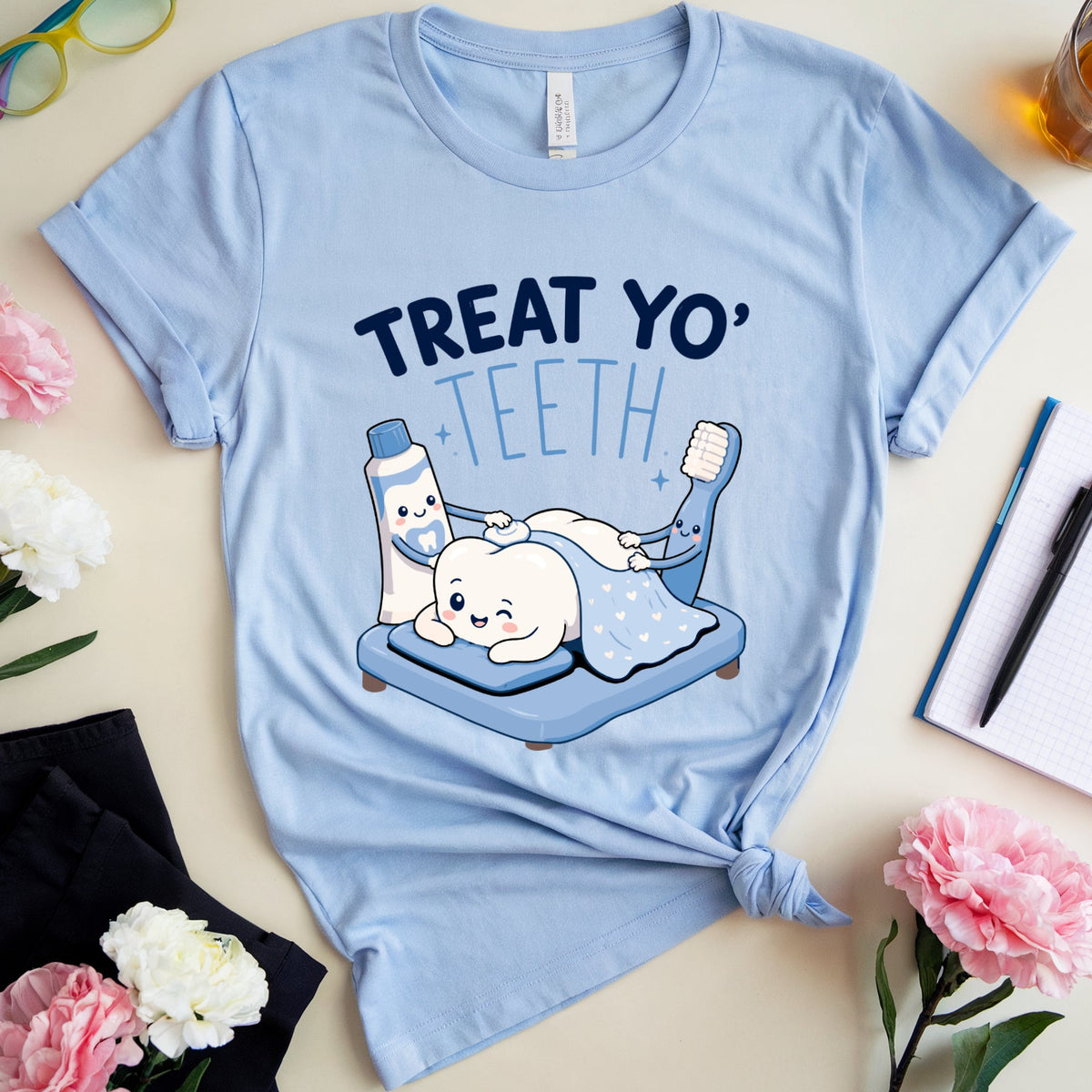 A light blue graphic t-shirt with a cartoon tooth illustration and the text 'TREAT YO' TEETH' is styled in a flat lay with pink and white flowers, eyeglasses, a notebook, and a drink on a light beige background.