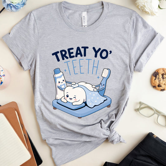 A heather gray graphic T-shirt laid flat on a light beige surface, styled with a tooth illustration and the words “TREAT YO' TEETH.” Surrounding props include a drink, notebook, pen, glasses, flower, and cookie.