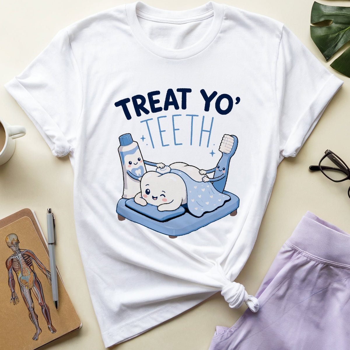 A white graphic t-shirt featuring a cartoon illustration of a toothbrush and toothpaste character treating a baby tooth, with the text 'TREAT YO' TEETH' printed above. The t-shirt is styled in a flat-lay arrangement on a beige surface alongside a cup of coffee, a notebook, eyeglasses, and lavender shorts.