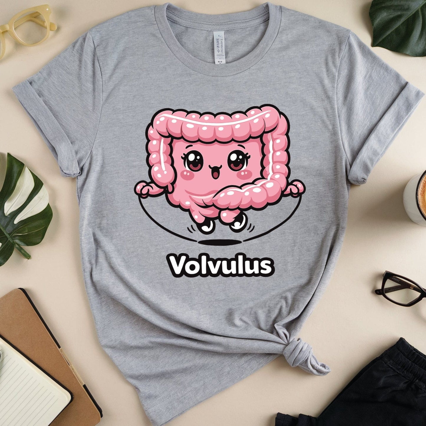 Gray t-shirt with a pink cartoon intestines design and 'Volvulus' text on a table with plants and a coffee cup.