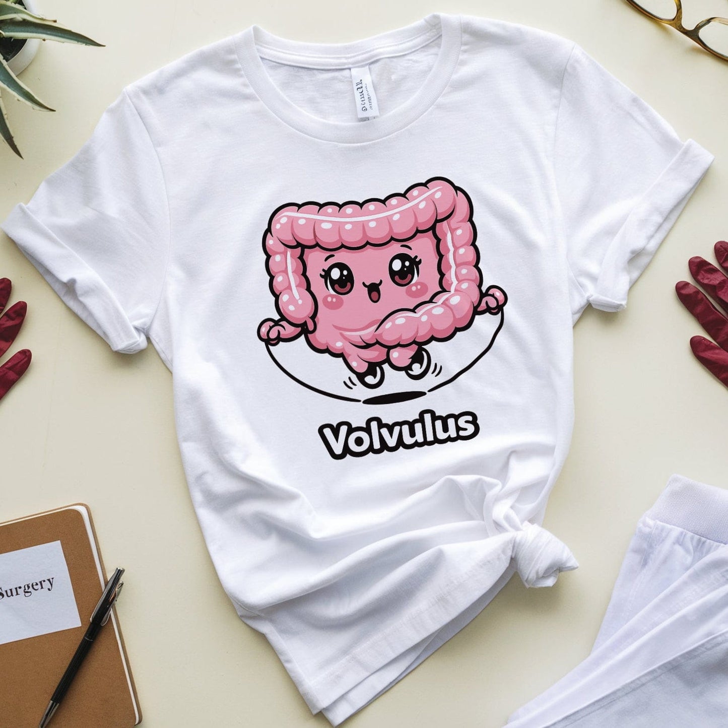 White t-shirt with a pink cartoon character and 'Volvulus' text on a light green surface with gloves and a plant.