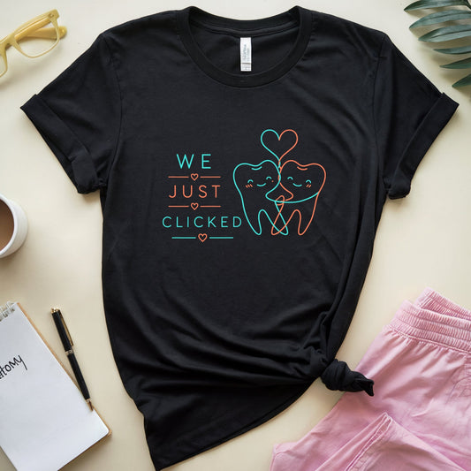 A black graphic t-shirt featuring a cute cartoon tooth design with the text 'WE JUST CLICKED' in teal and orange. The shirt is styled in a flat-lay arrangement on a light beige background with pink pants, a coffee cup, a notebook, and decorative leaves.