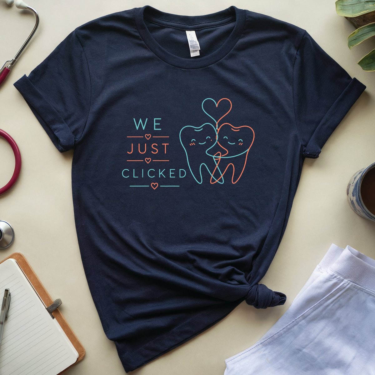 A navy blue graphic t-shirt with the quote 'lead us not into temptation to cancel the case.' in white text, styled in a flat lay with a stethoscope, notebook, plant, and coffee cup on a beige background.