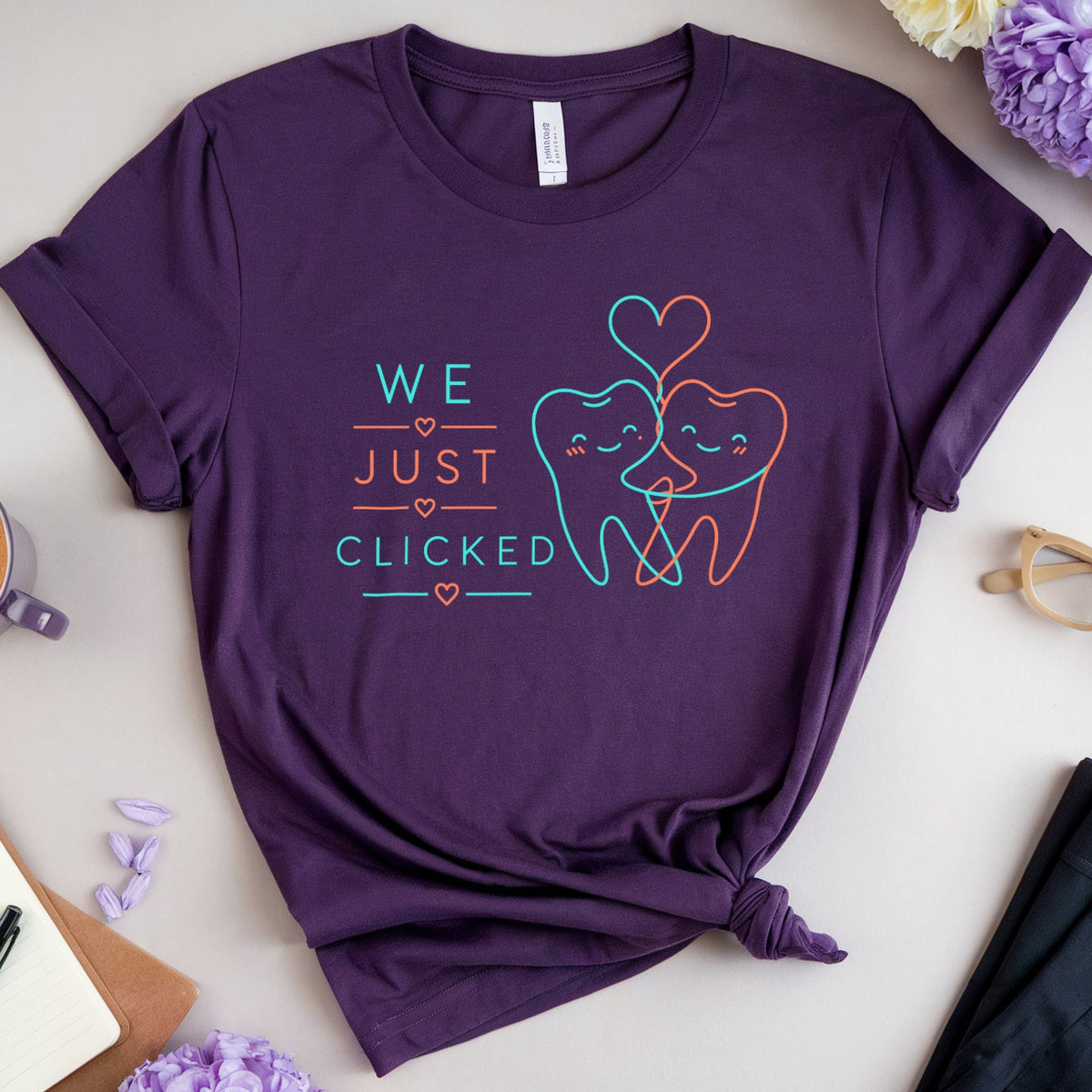 A purple graphic t-shirt with a 'WE JUST CLICKED' dental theme, styled in a flat lay with a coffee cup, notebook, glasses, and flowers on a light beige background.