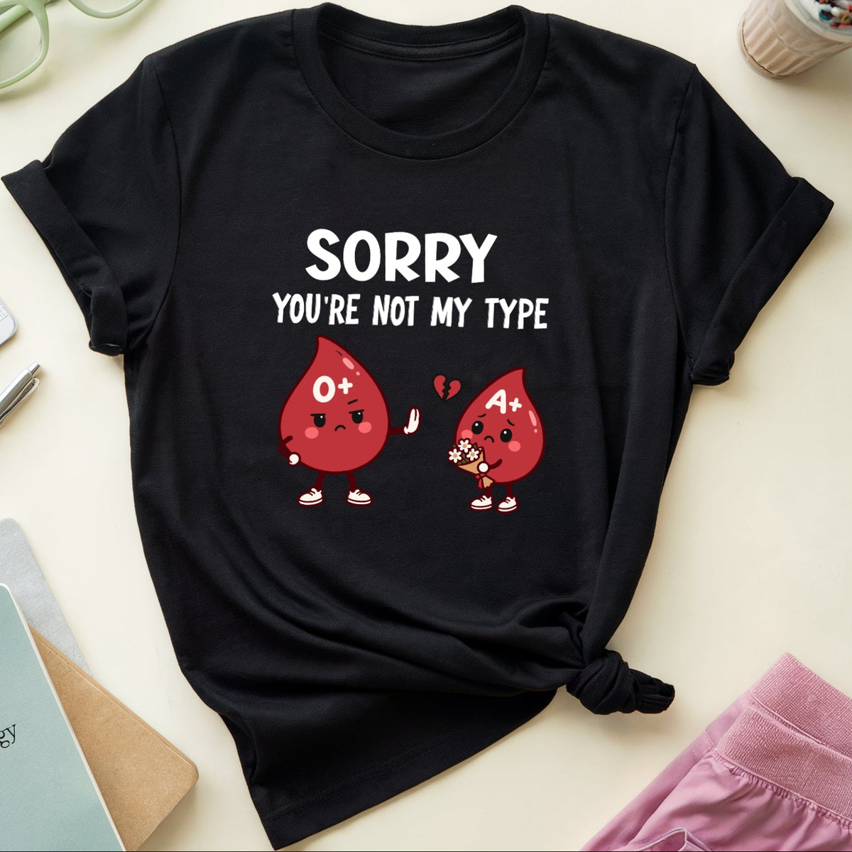 A black graphic t-shirt laid flat on a light beige surface, surrounded by props including eyeglasses, a drink, notebooks, and pink pants. The shirt features a white text print reading 'SORRY YOU'RE NOT MY TYPE' above two red blood droplet characters with '0' and '4' printed on them.