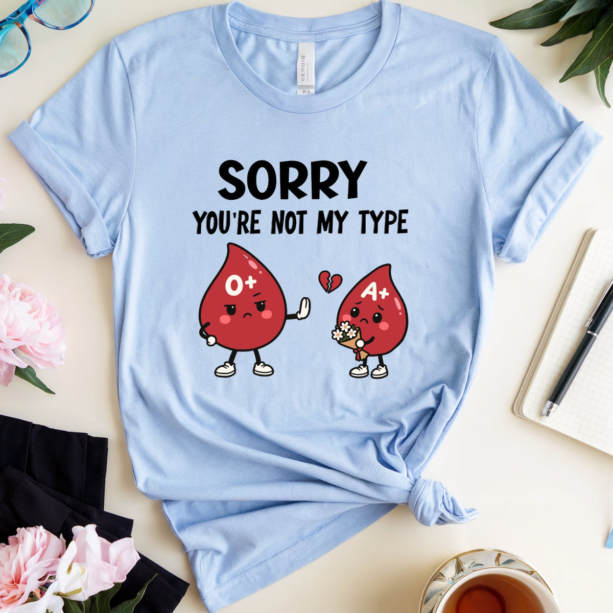 A light blue graphic t-shirt laid flat on a light beige surface, styled with pink flowers, sunglasses, a notebook, a pen, and a cup of tea. The t-shirt features a black text graphic reading 'SORRY YOU'RE NOT MY TYPE' above two cartoon red blood drop characters labeled O+ and A+ with a small red heart between them.