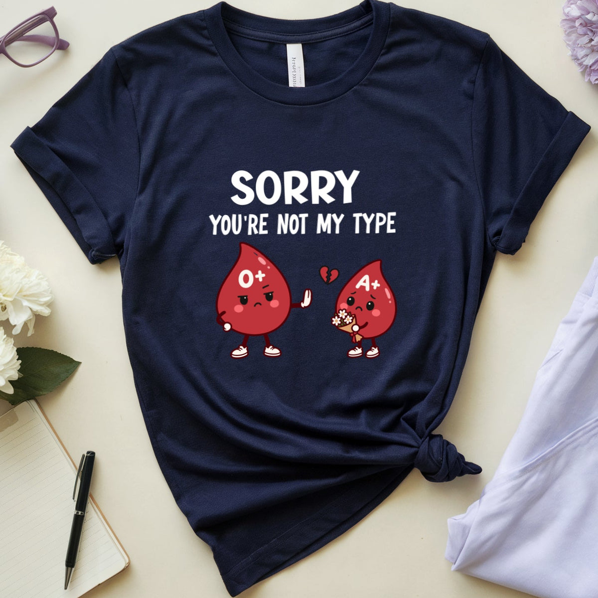 A navy blue graphic t-shirt is displayed in a flat-lay arrangement on a light beige background, styled with purple flowers, eyeglasses, a notebook, a pen, a cup of coffee, and light purple sweatpants. The t-shirt features a white graphic that reads 'SORRY YOU'RE NOT MY TYPE' with two red teardrop characters with '0°' and '40°' markings.
