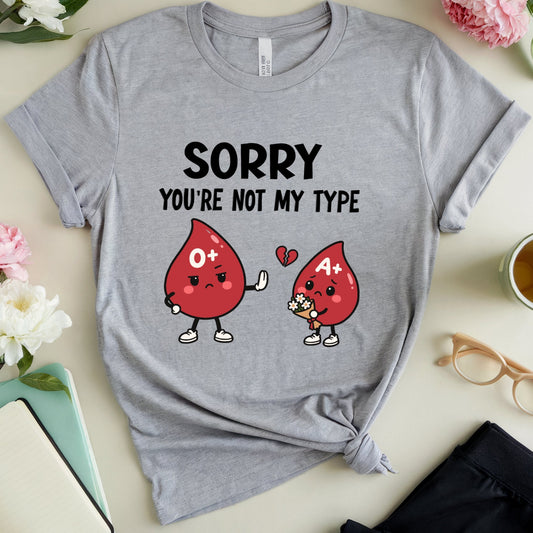 A heather gray graphic t-shirt laid flat on a light beige surface, surrounded by flowers, glasses, a notebook, and a cup of tea. The t-shirt features a cartoon graphic of two blood drop characters in O+ and A+ blood types with the text 'SORRY YOU'RE NOT MY TYPE'.