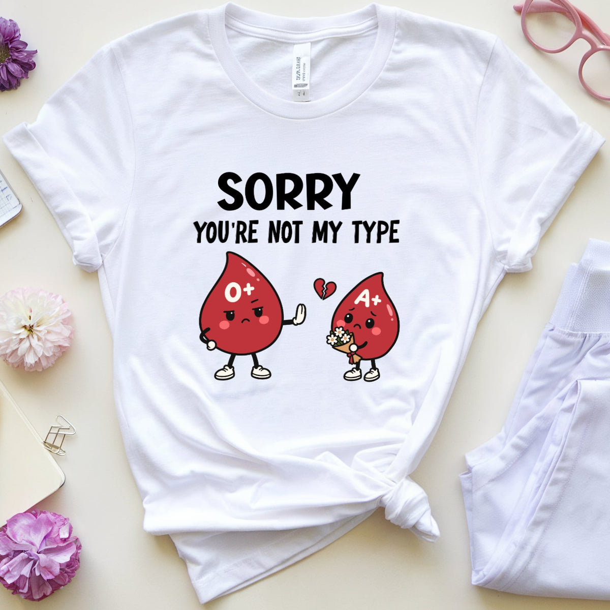 A white graphic t-shirt laid flat on a light beige background, styled with a knotted hem. The shirt features a cartoon graphic of two red blood droplets with hearts and the text 'SORRY YOU'RE NOT MY TYPE'.