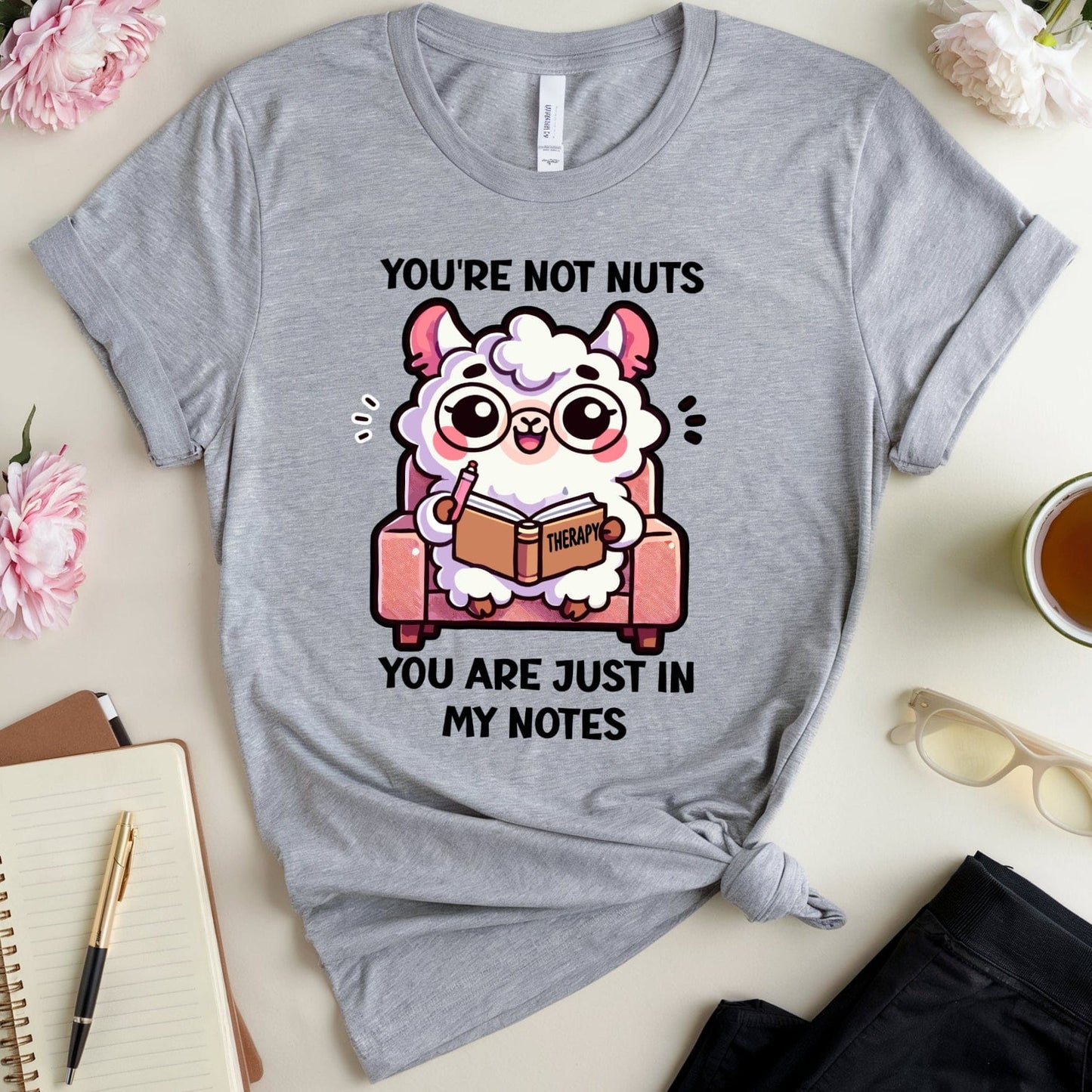 Gray t-shirt with cartoon llama graphic and text on a table with flowers and notebook.