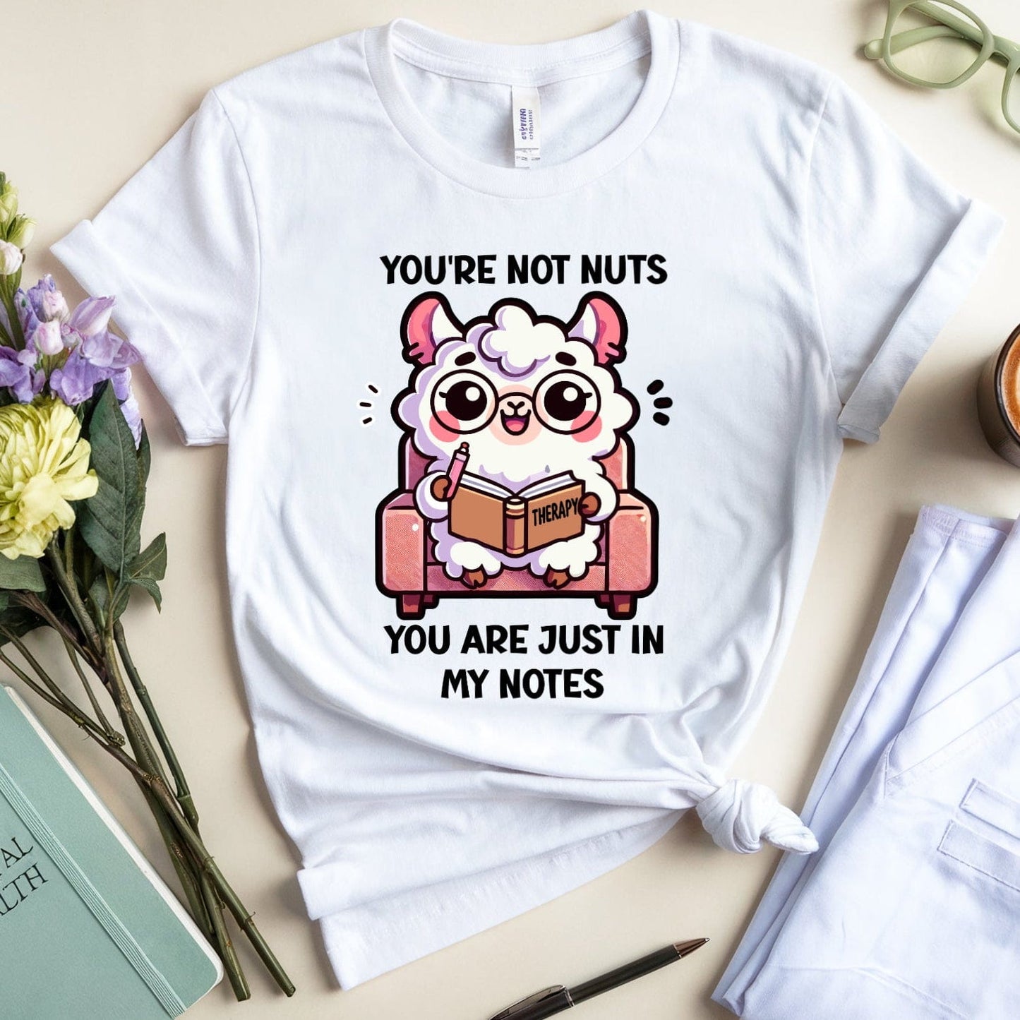 White t-shirt with cartoon llama graphic and text, placed on a surface with flowers, notebook, and coffee.