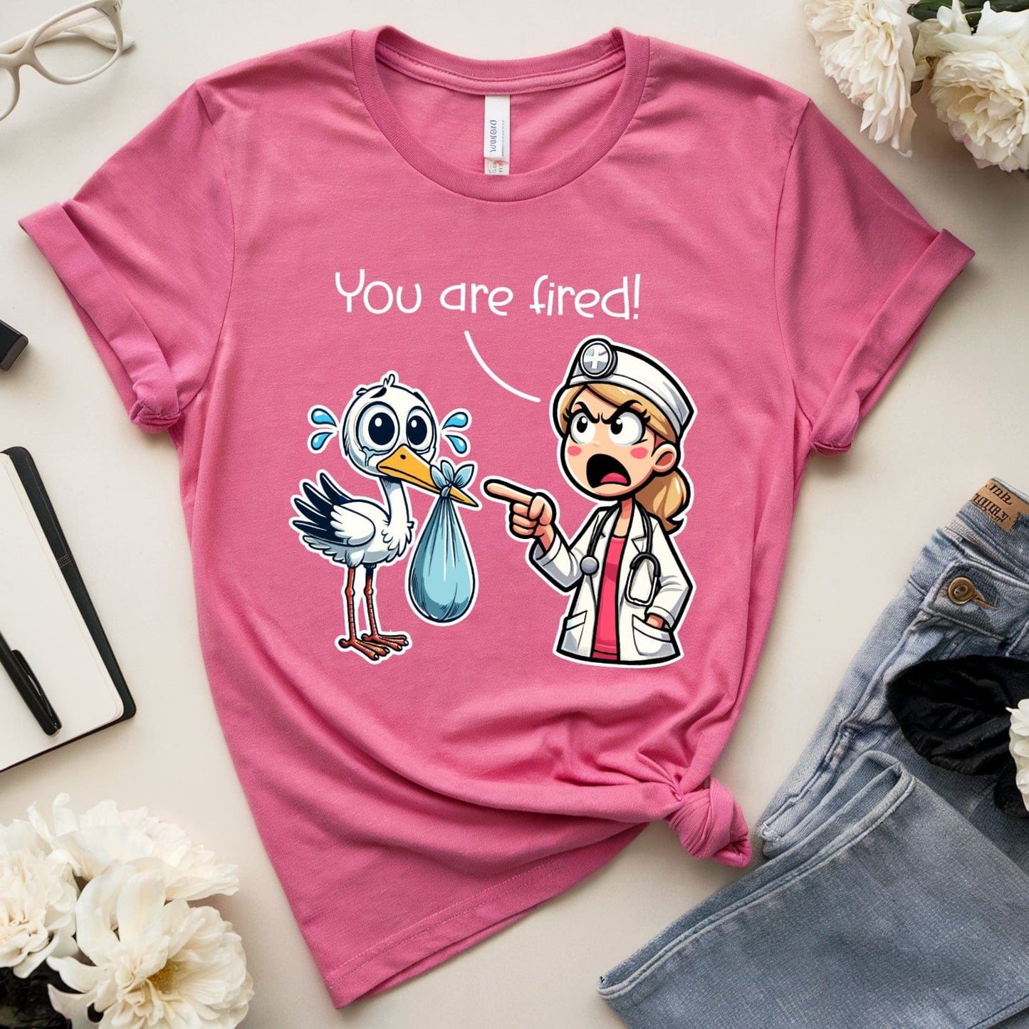 Pink t-shirt with cartoon design and text 'You are fired!' on a table with accessories.