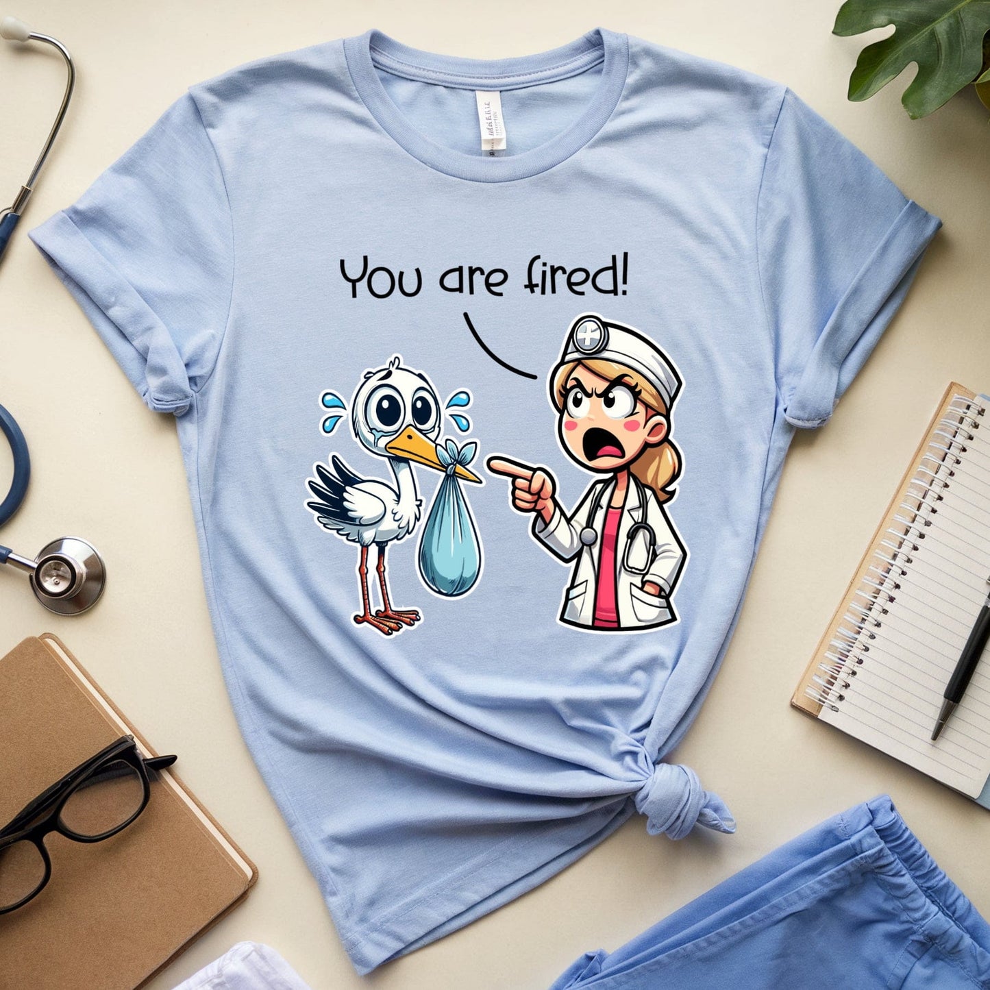 Light blue t-shirt with cartoon graphic and text 'You are fired!' on a desk with medical-themed items.