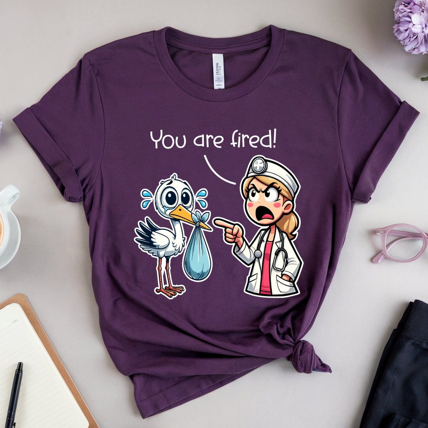 Purple t-shirt with cartoon design and text 'You are fired!' on a table with coffee and flowers.