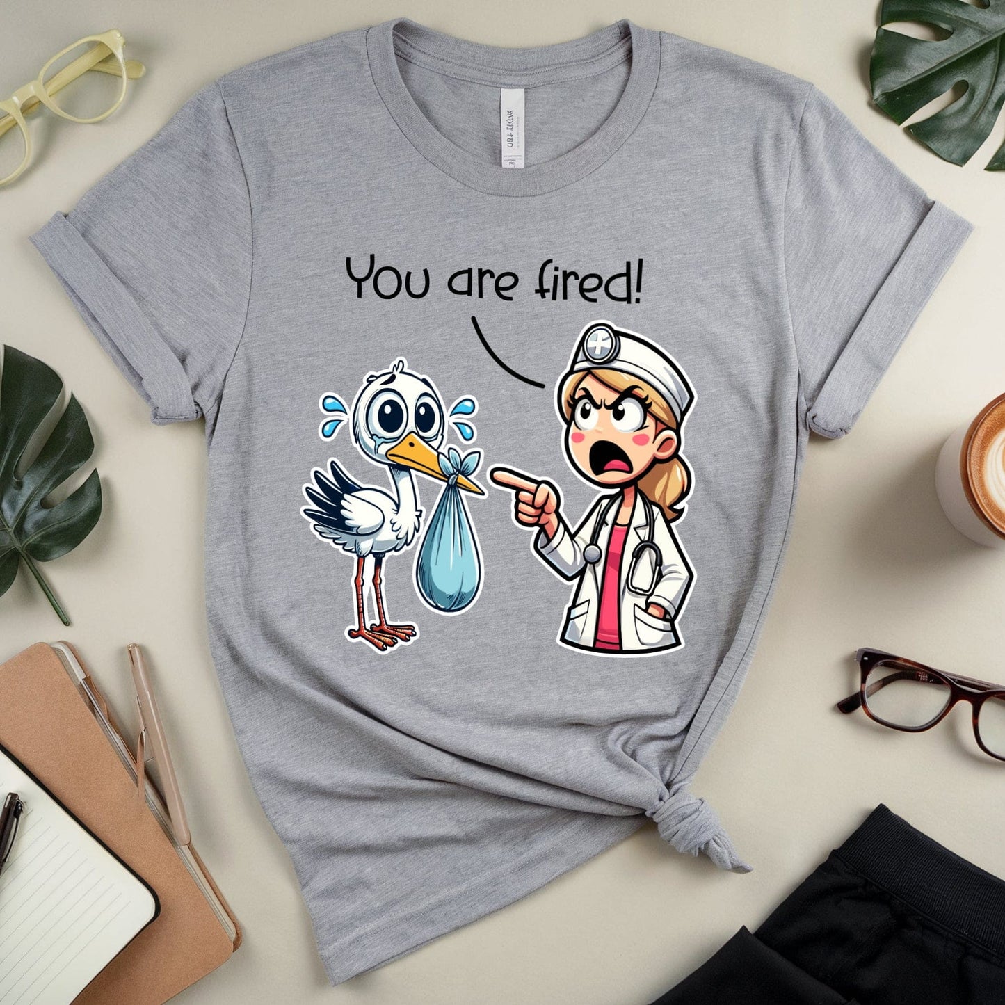 Gray t-shirt with cartoon design and text 'You are fired!' on a desk with office supplies.