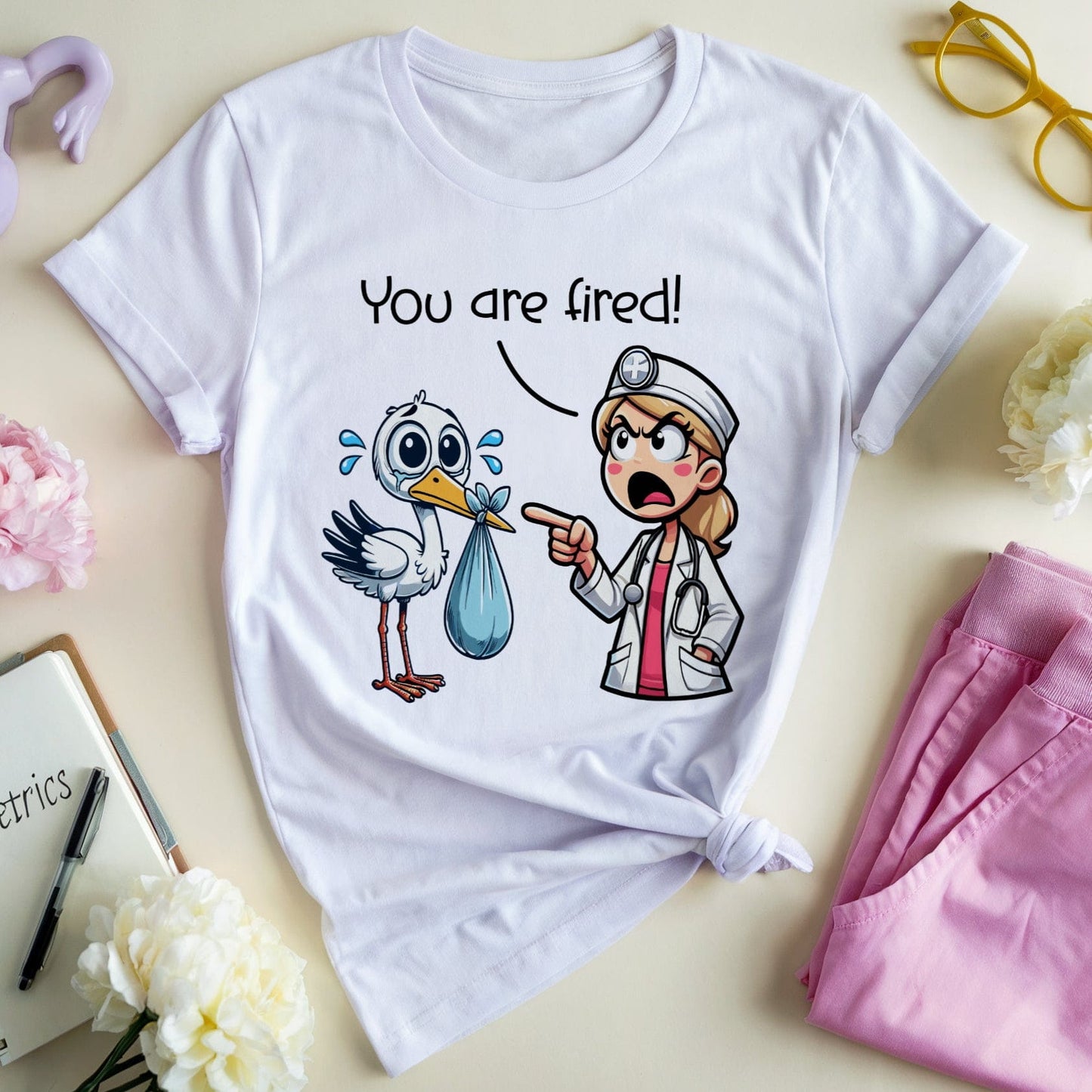 White t-shirt with cartoon design and text 'You are fired!' on a light background with flowers and a notebook.