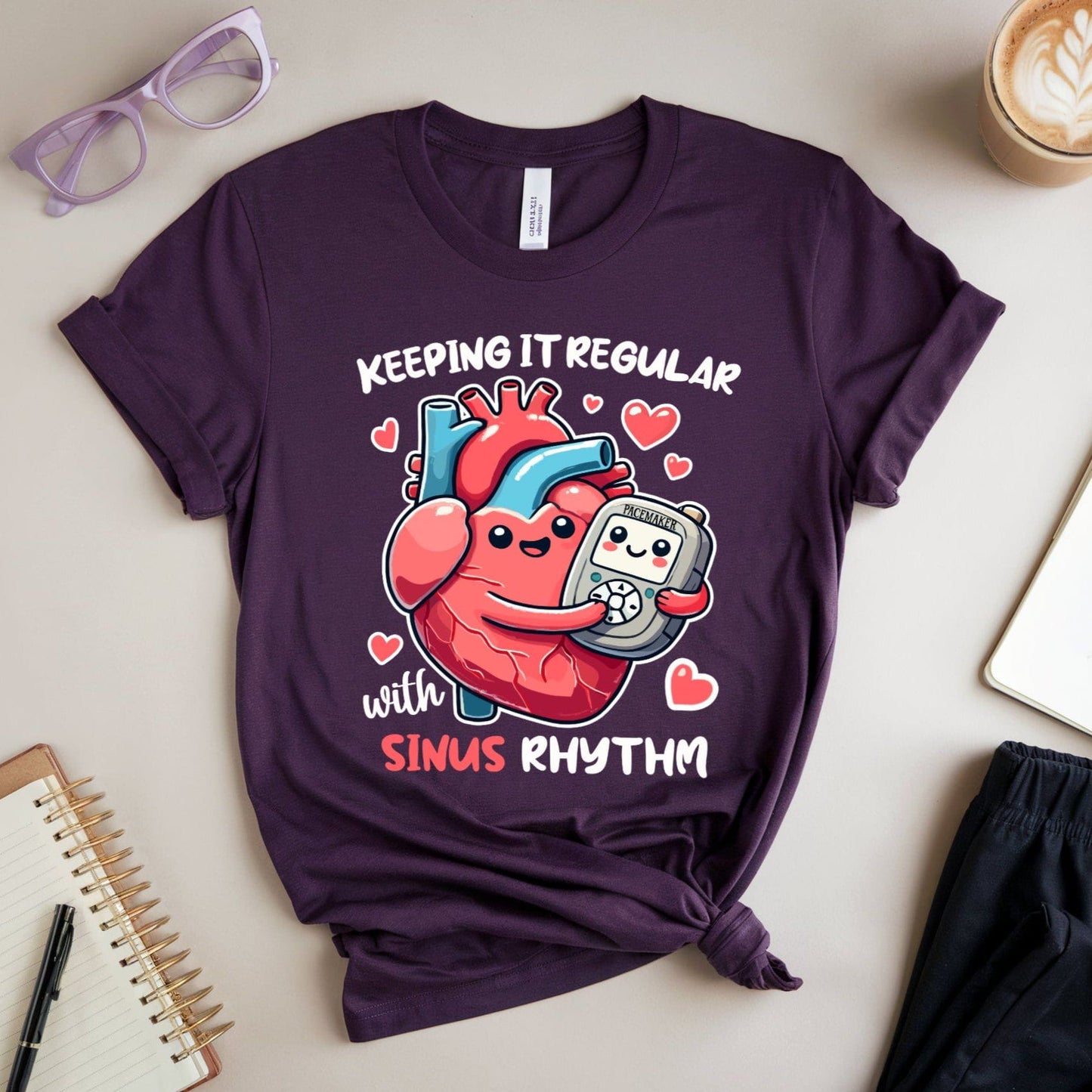 Purple t-shirt with a heart and EKG design, text 'Keeping it regular with sinus rhythm',  for cardiology on a light surface with accessories.