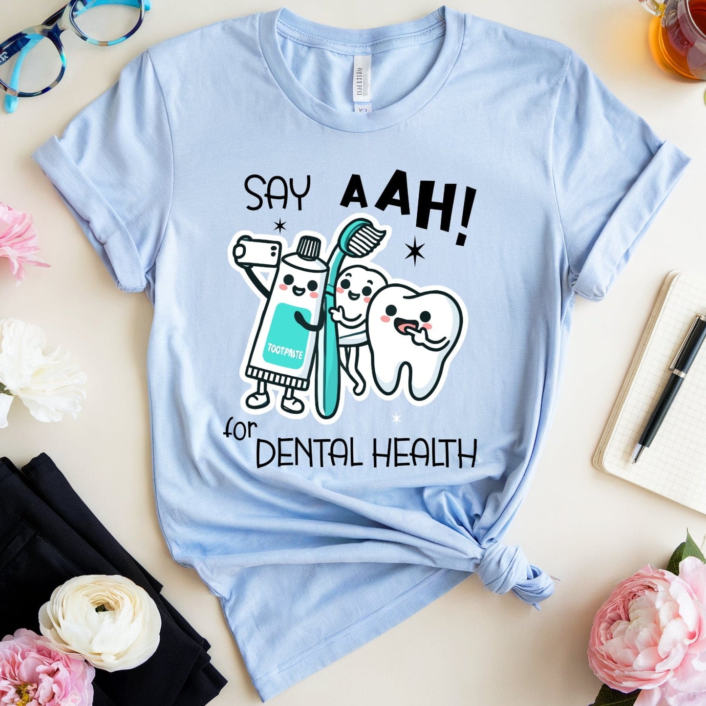 Light blue t-shirt with dental-themed design on a table with flowers and glasses.